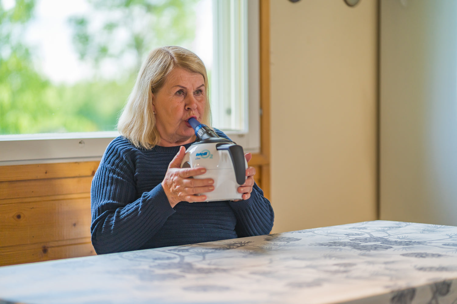 Woman is strengthening her lungs by blowing into a WellO2 steam breathing trainer in a room with a window and wooden furniture.